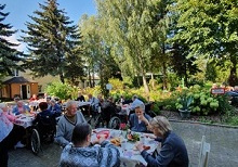 Zdjęcie przedstawia mieszkańców, którzy siedzą przy stolikach na świeżym powietrzu i jedzą smaczne kiełbaski z grilla