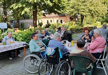 Zdjęcie przedstawia mieszkańców, którzy siedzą przy stolikach na świeżym powietrzu i jedzą smaczne kiełbaski z grilla . W tle widać piękny i zielony ogród.