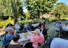 Zdjęcie przedstawia mieszkańców siedzących przy stołach w trakcie grilla , w tle widać piękny ogród