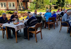 Mieszkańcy siedzący przy stołach na zewnątrz w trakcie grilla, w tle widoczny nasz ogród.