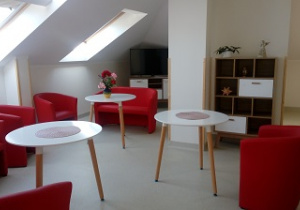 Zdjęcie przedstawia salę dziennego pobytu, na której widnieją białe stoły z czerwonymi fotelami, półkę oraz telewizor