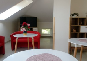 Zdjęcie przedstawia salę dziennego pobytu, na której widnieją białe stoły z czerwonymi fotelami, półkę oraz telewizor